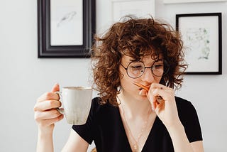 A curly, short-haired woman holds a mug of coffee while looking serious.