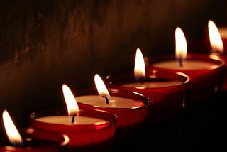 A close up photograph of a row of votive candles in auburn glass candleholders lined up along a concrete wall.