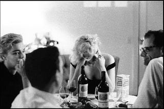 Black and white candid photograph of a dinner with four famous people deeply engaged in conversation around a small kitchen table: Arthur Miller, Marylin Monroe, Simone de Beauvoir, and actor Yves Montand.