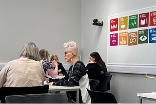 Two tables of women in discussion at a workshop. Both tables have three people at them. There is a display of the UN’s Sustainable Development Goals on the wall behind the tables.