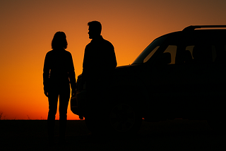 Image shows the silhouettes of a man and a woman standing at the front end of an SUV.