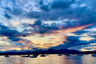 A blue sky littered with clouds floats over a pale ocean with boats in a quiet bay