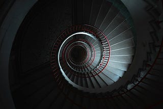 A spiral staircase with dramatic lighting, very dark and shadowy and the bottom isn’t visible