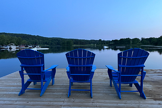 Three blue Muskoka chairs on a dock, facing the lake at dusk.