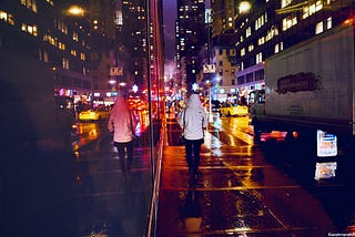 Person walking away from camera along a sidewalk on a busy street at night colour photo