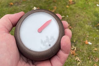 A hand holding the Homing Compass; a round, beautiful, sturdy looking device, which shows a single red arrow against a white background. The compass is designed to always points homeward.