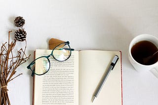 open diary with a pen and pair of glasses on it next to a mug of cofee and dried flowers.