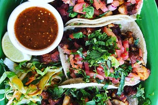 An overhead shot of a platter of soft tacos, with a ramekin of sauce and a lime wedge, on a green plate.