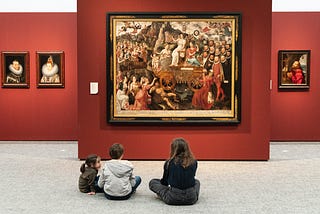 An adult and two small children sitting on the floor of a museum looking up at Tudor-era paintings hung on a deep red wall.