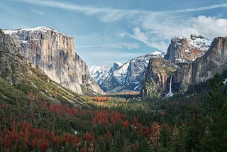 Yosemite National Park med den ikoniske El Capitan til venstre og Bridalveil Fall til høyre. Snødekte fjelltopper kan sees i bakgrunnen, mens forgrunnen viser trær med høstfarger som skaper en kontrast mot det storslåtte fjellandskapet. Det er en klar himmel med noen spredte skyer.