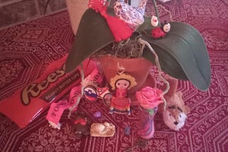A potted orchid on a red patterned rug surrounded by small objects, including a Reese’s Peanut Butter Cups pencil case, a red Chinese tassel, a blue-and-white bird figurine, a ladybug decoration, several pins, a small doll, a pink rose ornament, a miniature bouquet, a stuffed bird, and other small keepsakes arranged around the base of the plant.