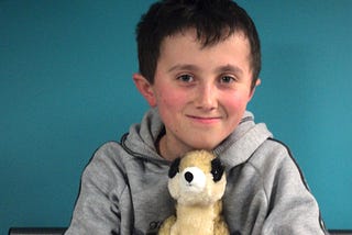 A young boy around 9 or 10 years old. He has brown hair and a small fringe. He’s smiling and sitting against a blue wall. He’s holding a stuffed animal of a meerkat. His cheeks are a little rosy, and is also wearing a grey sweater.