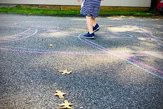 Image shows a blacktop with a pair of purple and blue chalk lines twisting throughout. A small child’s feet walk along the lines.