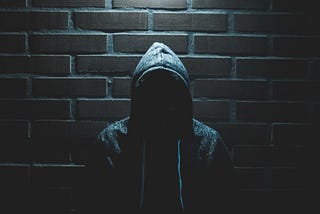 Image of a hooded man, face not visible, stood against a brick wall, beneath a light