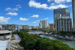 Photo of the view from a Honolulu hotel window; deep blue sky, tall buildings on the right, green trees and water in the foreground