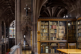 A neo-Gothic style library. Upstairs with a hallway and arched walls and pillars. On the right side, a book-case of rare books and a wooden table on the bottom right with two chairs.