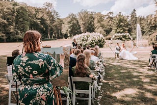 Photo of me in a green floral dress, behind the seated guests. During outdoor wedding ceremony. Standing, painting.
