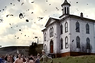 Color still from the film, “The Birds,” showing children and actress Tippi Hedren fleeing from birds swarming in the sky with a building in the background.