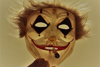 A mask of a crazy clown face and wild hair with a cigarette sticking out of its mouth is held up by a hand, partially in view