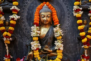 Gilded statue of the Buddha against a dark grey, carved backdrop and draped with garlands of orange, red, and yellow flowers.