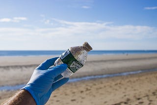 A person’s left hand in a protective rubber glove holding up an empty, dirty single-use plastic water bottle in front of a pristine beach, presumably there collecting trash.