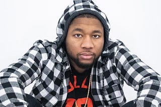 Adrian Prince seated with his elbows resting on his raised knees as he looks directly into the camera in a close-up head shot. Adrian Prince is wearing a black and white plaid pullovers with a gold necklace laying atop the black t-shirt with red writing that he’s wearing beneath his plaid pullover.