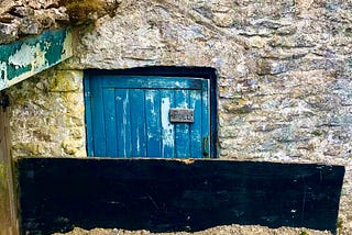 A small door nestled within a stone wall. There is a small sign which reads: “Pull”.