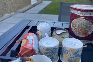 A tea tray on a garden table, with someoe pouring tea into a cup while two people are walking at the back of the garden. They look blurred.