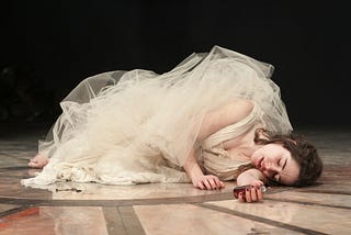 A young woman dressed in white lays on the ground as if deceased.