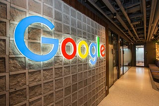 Illuminated Google logo in blue, red, yellow, and green on a textured stone wall in a modern office hallway with exposed ceiling.
