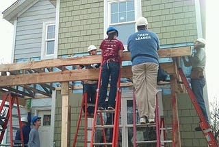 People with construction helmets stand on ladders, constructing a porch roof in front of a nearly completed Habitat for Humanity house. One is the author’s younger daughter. (Photo by author.)