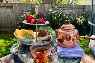 A tiered tea tray with a egg and cheese sandwich on a sourdough pretzel bun, corn chips and guacamole, and strawberries and dark chocolate on the top tier. Next to it is a pink glass cup filled with black tea and a copper teapot. It all sits on a small garden table outside.