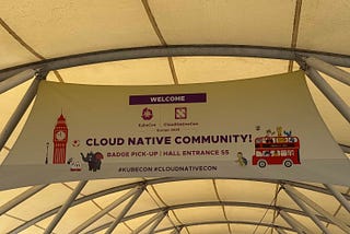 A banner hanging under a tent structure welcomes attendees to KubeCon + CloudNativeCon Europe 2025. The banner features the text “WELCOME” followed by the logos of KubeCon and CloudNativeCon. Below, it reads “CLOUD NATIVE COMMUNITY! BADGE PICK-UP | HALL ENTRANCE S5” and includes the hashtags “#KUBECON #CLOUDNATIVECON”. The banner is decorated with illustrations of Big Ben, an elephant, a zebra, a red double-decker bus filled with various animals, and a turtle.