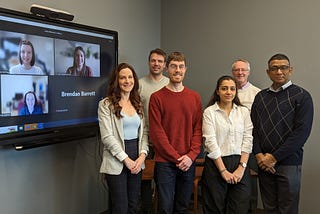 A group photo of nine people smiling. Three people are smiling on a tv screen from a virtual platform.