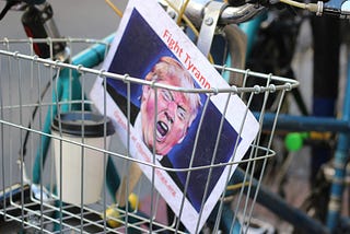 Photograph of a wire basket of a bicycle containing a to-go cup of coffee and a protest placard showing Donald Trump shouting with a reddened face with “Fight Tyranny” written in red at the top. (Photo by Samantha Sophia for Unsplash.)