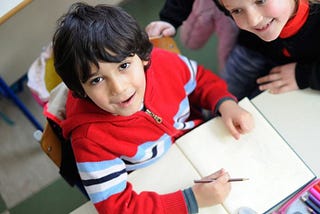 A small boy is sitting in front of a notebook with a pencil in his hand. He looks up at the camera with a smile. The girl next to him smiles at him admiringly.