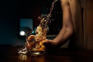 Against a dark background, a woman in a sleeveless, tan top slams a glass of whiskey down hard on a mahogany table, sloshing liquid everywhere.