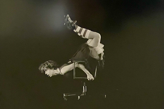 A black and white photo of a short-haired dancer rolling across a stage upside-down on an office chair. She is wearing calf-high, horizontally striped socks.