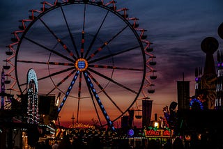 A Ferris wheel can be seen looming over the fairgrounds of a carnival, backdropped by a sky just at the end of sunset, with a gradient from dark blue at the top of the image, to purple and then red.