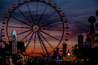 A Ferris wheel can be seen looming over the fairgrounds of a carnival, backdropped by a sky just at the end of sunset, with a gradient from dark blue at the top of the image, to purple and then red.