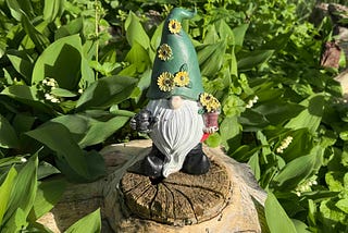 A garden gnome with a green hat decorated with yellow flowers stands atop a tree stump, surrounded by vibrant greenery. The gnome has a long white beard, black boots, and holds a small watering can in one hand and a flower pot in the other. Sunlight highlights the lush foliage and the intricate details of the gnome’s features.
