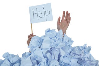 A woman’s hand rises from a pile of crumpled paper. Her other hand is holding a sign that says “Help.”