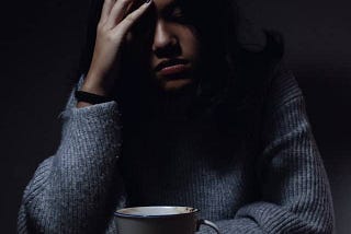 Depressed woman sitting in a dark room. Her hand rests on her forehead in obvious stress. A cup of coffee sits in from of her. She is wearing a sweater.