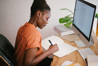An African girl sitting down and writing into her notepad in front of a Mac computer screen
