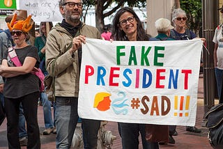 Rally attendees hold a banner containing a cartoon profile of Trump and  colorful letters in the style of a tweet, reading “FAKE PRESIDENT #SAD!!!”