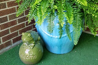 Bright green ceramic frog sitting on a large ceramic ball with the word “Welcome” imprinted on it, in front of a large turquoise pot of Boston ferns.