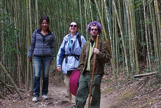 Three people walking through a bamboo forest in the mountains of Taiwan. One is a man wearing a green jacket and purple knitted hat, and the other two are women. All appear to be smiling or laughing.