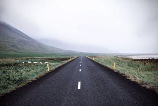 A two-lane road heads toward large hills and a cloudy sky.