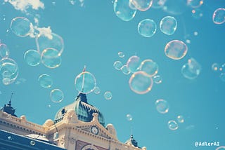 A photograph of the partially viewable top of a train station building. Bubbles are floating in the air against the blue sky above.