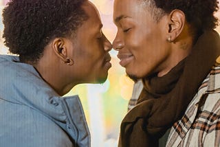 Two Black, masculine presenting people wearing coats are closing their eyes and touching their noses together in an intimate pose while holding hands.